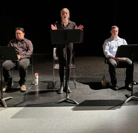 A young woman standing and reading from a music stand with two young men sitting on either side