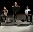 A young woman standing and reading from a music stand with two young men sitting on either side