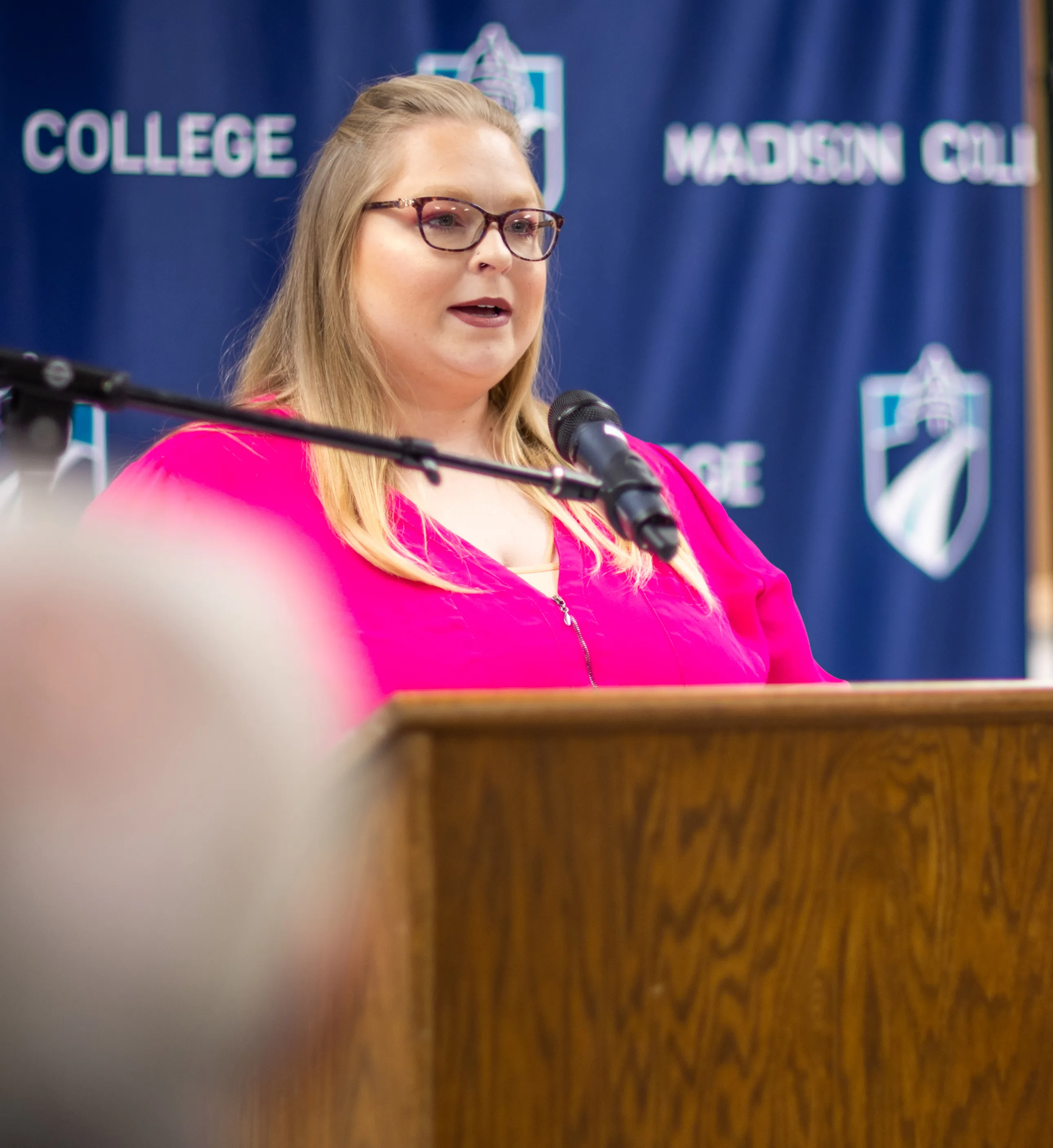 Woman (Natalie Wepking) with blond hair and pink shirt, standing at podium talking.