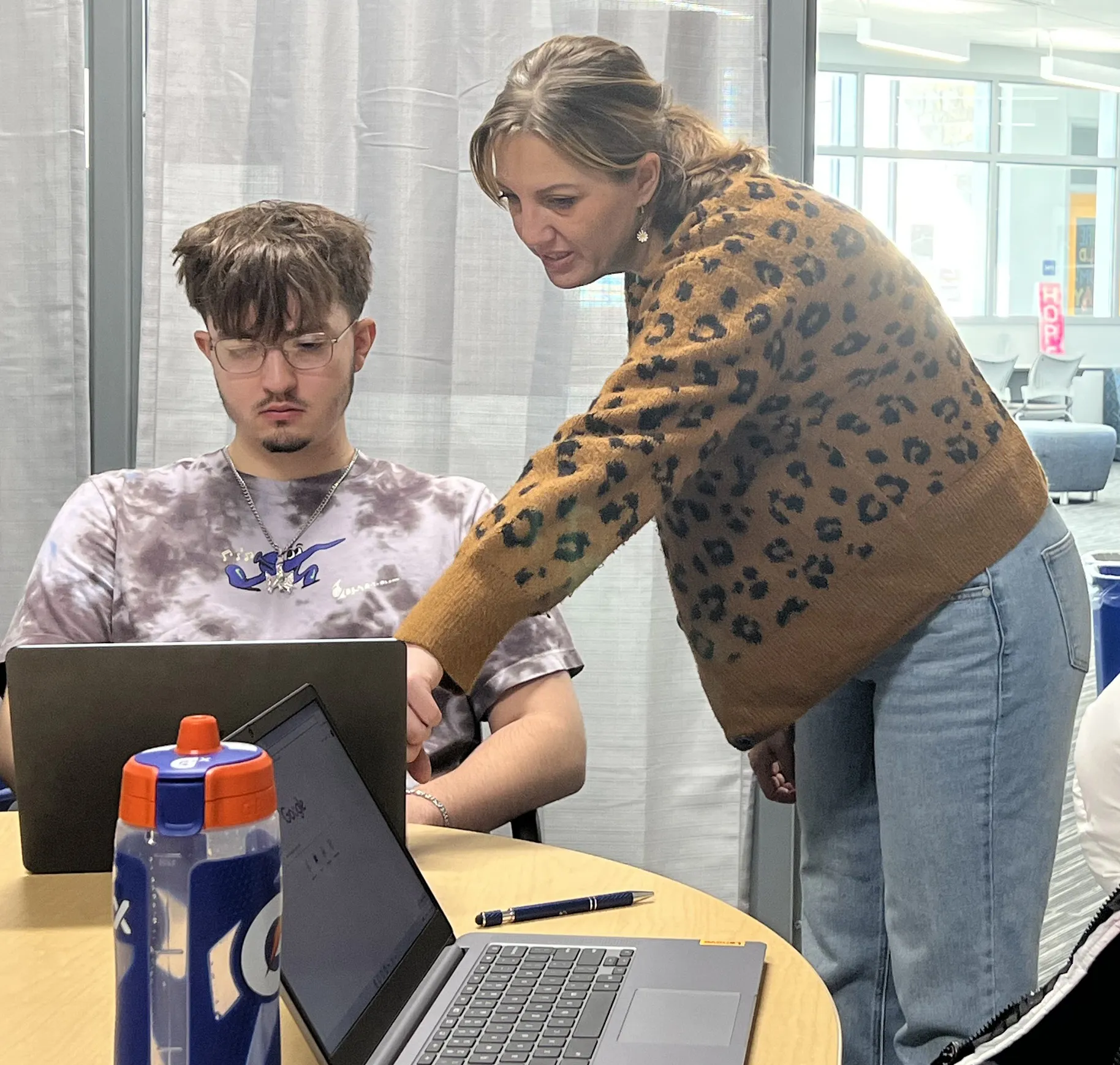 Madison College woman navigator, wearing brown sweater and jeans, leaning over a laptop pointing out something to a student, who is sitting on a chair.