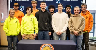 Group of apprentices, dressed in orange, yellow, black, white and brown sweatshirts, posing for a photo.