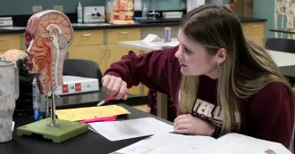 Female student, wearing dark red sweatshirt, sitting at point to an anatomy model. Text books on desk in front of her.