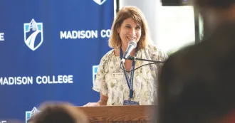 President Berne at a podium, talking into a microphone, while people look on.