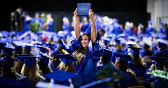 Graduate proudly holding up diploma over her head while fellow graduates sit next to her in the commencement ceremony.