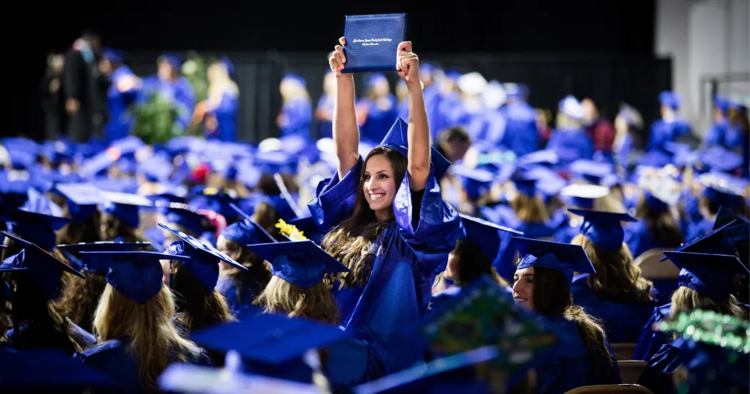 Graduate proudly holding up diploma over her head while fellow graduates sit next to her in the commencement ceremony.