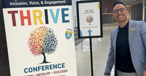 Madison College administrator smiling, wearing blue blazer, standing next to Thrive Conference placard.
