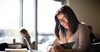 Female student sitting at desk, reading a book, and smiling.