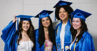 Four female grads, in blue cap and gowns, smiling and posing for camera.