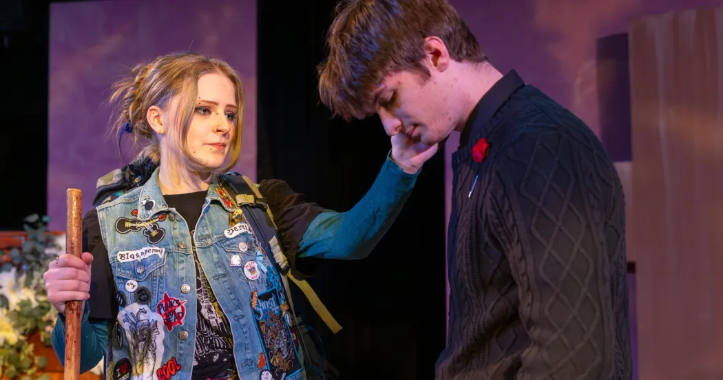 Woman actor with blonde hair, denim jacket with patches, touching the cheek of a man, who bowing his head and looking sad.