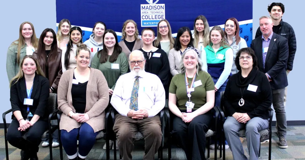 Group of people, students, faculty and administrators, sitting in chairs posing for a photo, with a Madison College logo in the background.