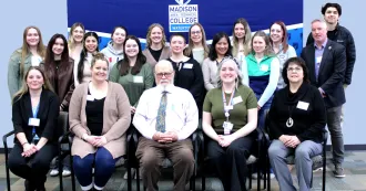 Group of people, students, faculty and administrators, sitting in chairs posing for a photo, with a Madison College logo in the background.