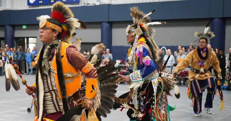Three Native American dancers in bright regalia with feathers. Audience in the background watching the powwow event.