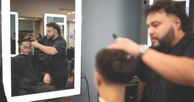 Student in barbering class cutting other person's hair, while standing in front of a mirror.