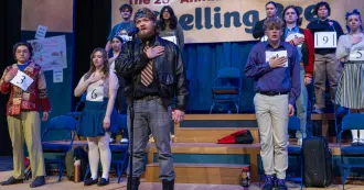 Group of 10 actors on stage of spelling bee musical, performing with hands at their heart, saying the Pledge of Allegiance.