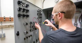 Male student with short dark blonde hair and black T-shirt, adjusting equipment in an electrical box with a screwdriver.