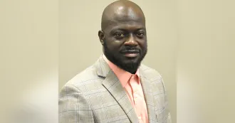 Man with dark skin, beard, light brown blazer and peach shirt.