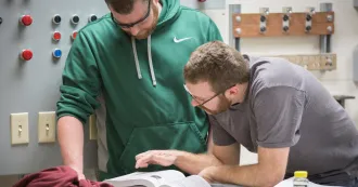 Two apprentices, looking at a text book, inside a classroom with gray electrical boxes on the wall.