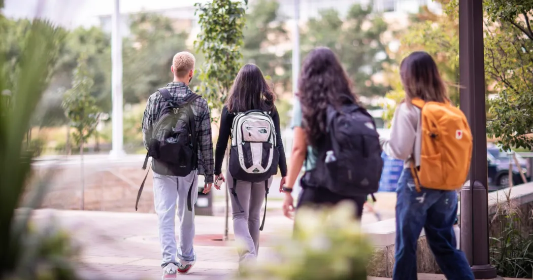 Four students, wearing backpacks and jeans, walking on campus.
