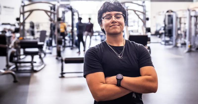 Student with black shirt, dark hair, hands crossed, smiling in a background of gym, with equipment in the background.
