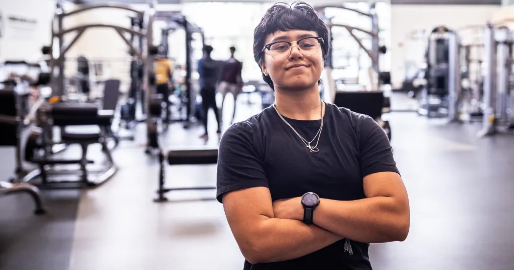 Student with black shirt, dark hair, hands crossed, smiling in a background of gym, with equipment in the background.