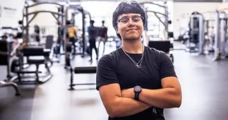 Student with black shirt, dark hair, hands crossed, smiling in a background of gym, with equipment in the background.