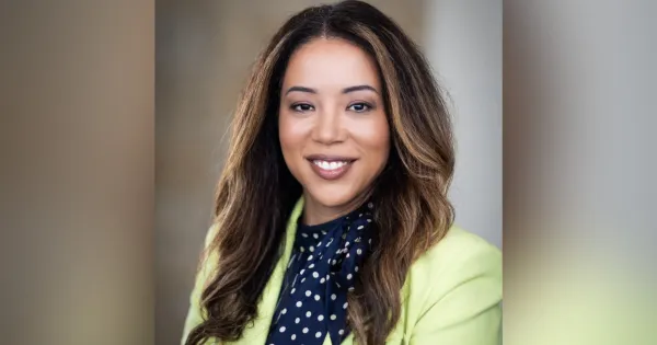 Woman with brown long hair, blue blouse and yellow blazer, smiling at camera.