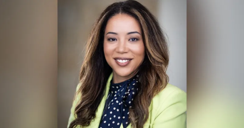 Woman with brown long hair, blue blouse and yellow blazer, smiling at camera.