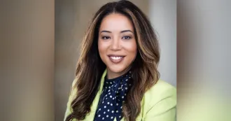 Woman with brown long hair, blue blouse and yellow blazer, smiling at camera.