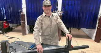 Man with brown shirt and brown baseball cap, in a welding lab, with a piece of steel in his hand.