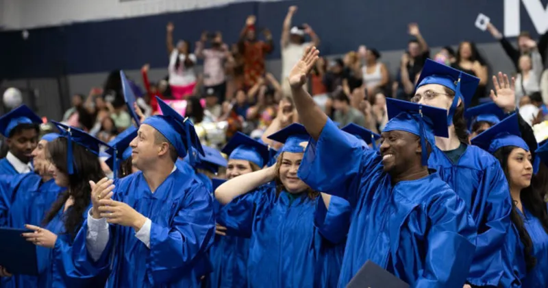 Graduates dressed in blue gowns and caps, smiling and waving to friends and family in a large ceremony setting.