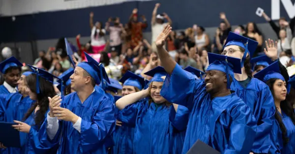 Graduates dressed in blue gowns and caps, smiling and waving to friends and family in a large ceremony setting.