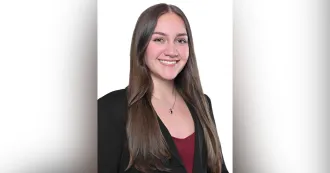 Woman with long brown hair, black blazer and red shirt, smiling.