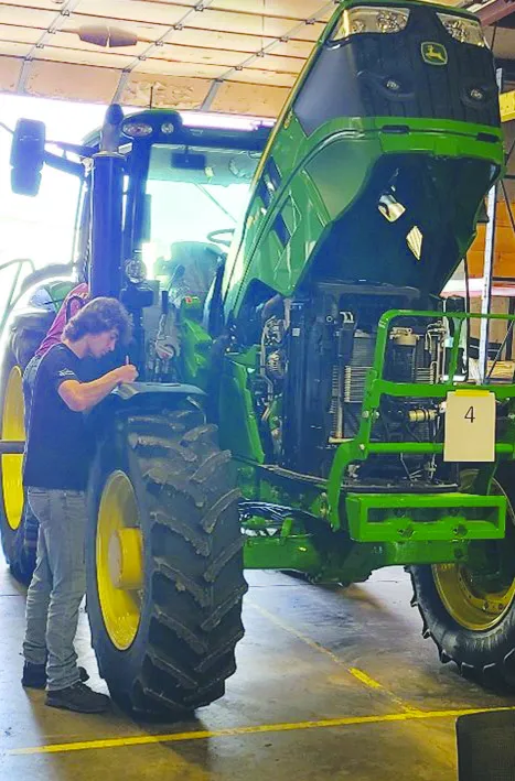 Male student, wearing black shirt and jeans, working on a green John Deere tractor inside an equipment bay.