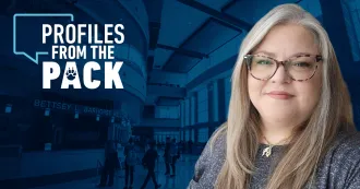 Image of Wendy Franczak, wearing a blue grey shirt, silver necklace, smiling at camera. Profiles from the Pack logo with wolf paw in background.