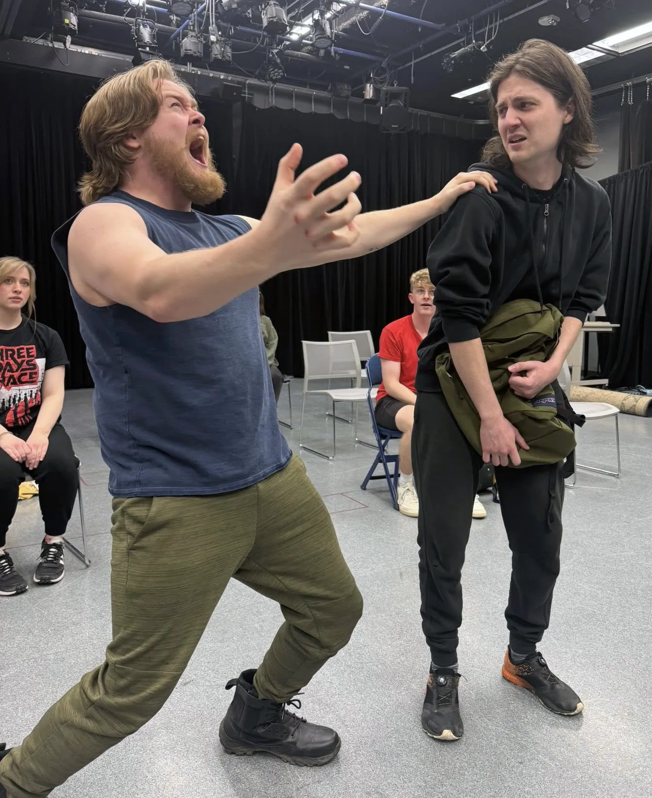 Male actor, wearing a blue shirt and green pants, holding hands out and singing, while another male actor, wearing black pants and black shirt reacts.