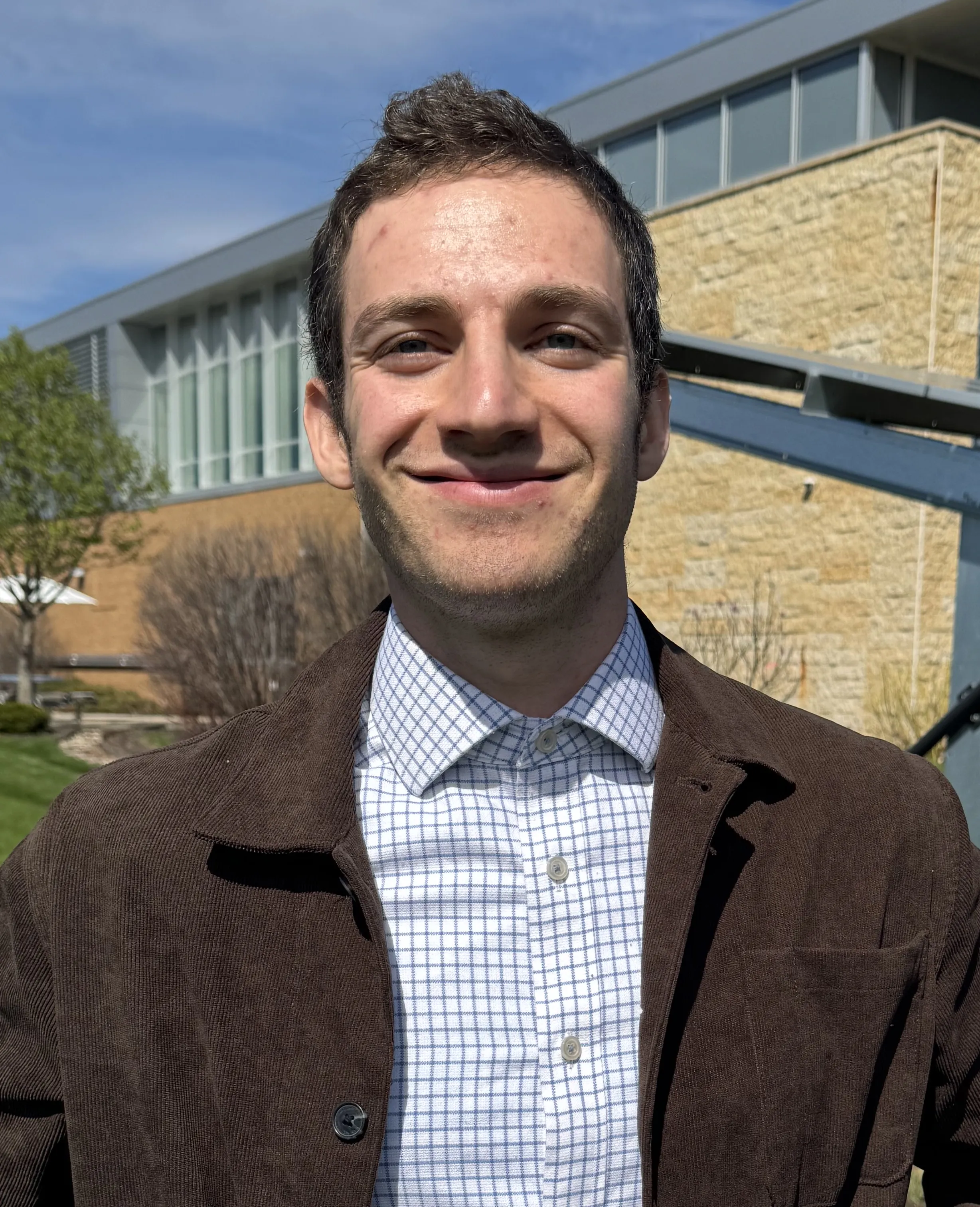 Male with short dark hair, blue check shirt, brown blazer, smiling, standing in front of Truax campus building.
