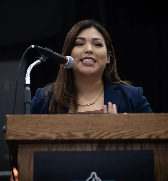 Woman with dark hair and a blue blazer speaking at a podium with a microphone in front of her.