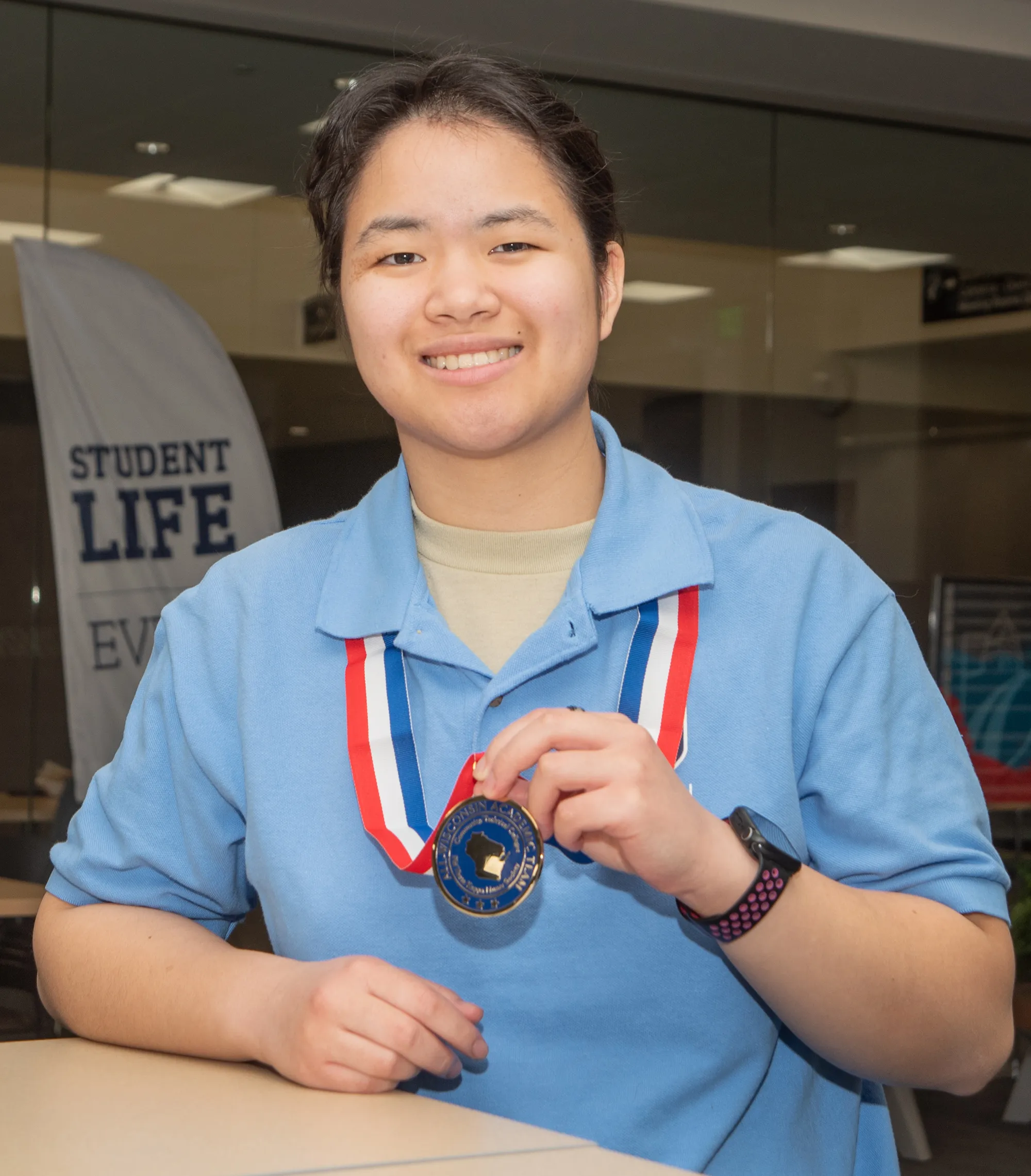 Student with dark hair, light blue shirt, wearing a medal with a red, white and blue ribbon.