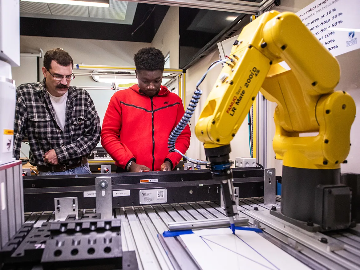 faculty and student standing by a yellow robot arm, the arm is drawing. 